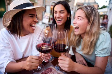Three cheerful female friends toasting with glasses of red wine, laughing and savoring their time together at an outdoor restaurant, enjoying the vibrant atmosphere of summer