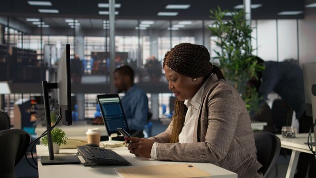 African american manager takes a break from business work and browsing social media, checks messages and websites at her desk. Analyst multitasking and texting during schedule. Camera B. - Powered by Adobe