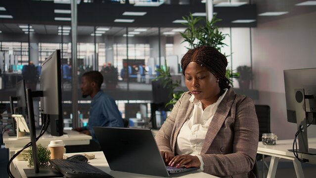 Confident businesswoman at her office desk working on report writing and answering emails, administrative tasks management. Black female expert reviewing objectives to ensure success. Camera B.