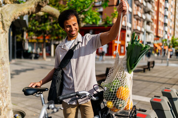 Happy businessman is holding a reusable shopping bag with groceries next to his bicycle, promoting...