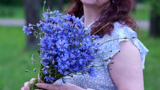 A woman's shoulder and arm visible as she turns to the camera and offers a bouquet of cornflowers. A gentle gesture of greeting or gift. 4K romantic nature footage.