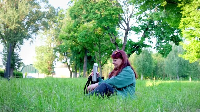 Woman sitting on grass in a sunny park, pouring hot tea from a thermos and sipping from a cup. Peaceful outdoor moment in 4K.