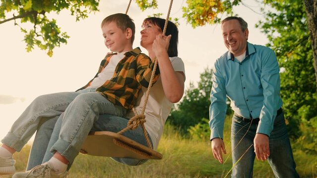 Happy family swinging on swing together at summer greenery tree park enjoy weekend. Smiling mother and cute boy son sitting father pushing playing relaxing sunny natural garden with positive emotion - Powered by Adobe