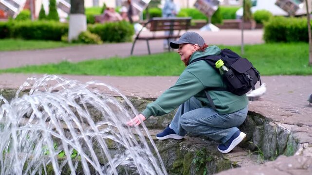 Woman touches fountain jets with hand and relaxes in city park. 4k video