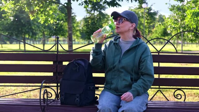 Sporty woman in sunglasses resting on park bench, taking water bottle from backpack and drinking. Healthy lifestyle and outdoor wellness in 4K.