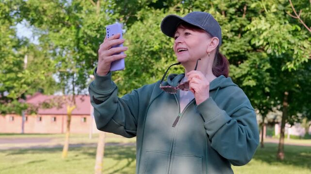 Sporty smiling woman in sunglasses and baseball cap flirting playfully at smartphone camera, enjoying sunny day. 4K lifestyle footage.