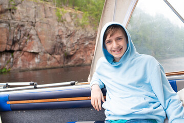 A 12 year old boy sits on a boat in front of a mountain shore on a water excursion.	