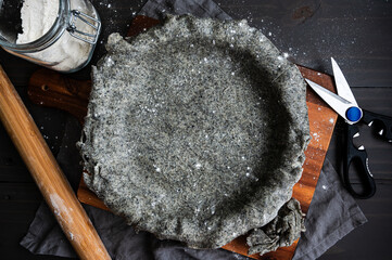 Black Sesame Pie Crust Draped Over a Pie Plate: Rolled out savory pie crust dough in a pie pan with a rolling pin, kitchen shears, and jar of flour