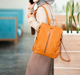 The body of a woman wearing a hijab and formal attire is making a call on her smartphone while carrying a large brown leather backpack hanging from her shoulder.