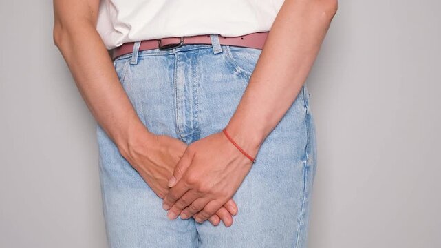 Close-up of a woman in jeans and white t-shirt standing with hands clasped in front of her lower abdomen, symbolizing the discomfort and urgency related to bladder leakage or urinary incontinence