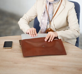 A woman's body wearing a hijab and formal attire takes a gadget from a brown leather bag on the table.
