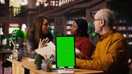 Mature colleagues team review paperwork next to chroma key mockup, using multiple academic sources and books in classic library setting. Learning space with library resources. Camera A.