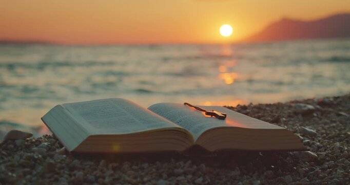 Open Bible book on pebbles with cross pendant during tranquil beach sunset