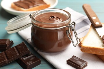Tasty chocolate butter in jar, pieces, knife and bread on light blue wooden table, closeup