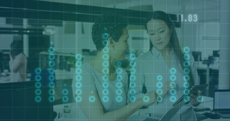 Two office professionals pointing tablet and viewing data in open-plan office, laptop data overlay