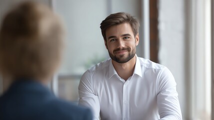 A young man in a white shirt smiles warmly while engaging in a conversation with a blurred person in a bright, professional indoor setting.