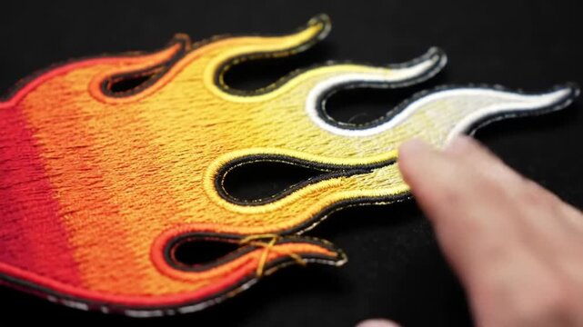 Close Up of Vibrant Flame Embroidered Patch with Detailed Textile and Hand Manipulation on a Dark Surface