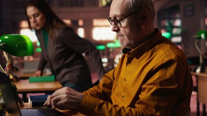 Senior male student reading educational papers on laptop in a university library, committed to completing coursework and achieving a meaningful degree in his later years. Old man. Camera B.
