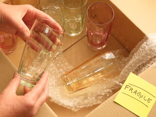 Closeup of woman hand holding a colored glass cup over a cardboard box with colored drinkware and...