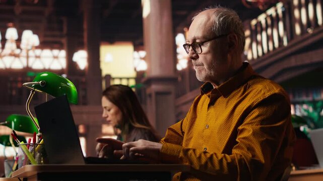 Focused senior guy reading academic text on laptop at the university, sitting in vintage study room and browsing online documents. Lifelong education through formal learning. Camera B.