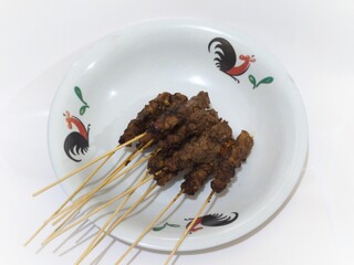 Ten skewers of beef satay are served on a ceramic plate with a classic rooster motif.
