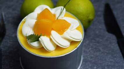 Small mango mousse cake decorated with whipped cream rosettes, mango cubes, and mint leaves, sitting on a dark gray textured surface with two limes and a knife in the background