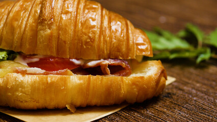 Freshly made croissant sandwich filled with crispy bacon, lettuce, tomato, and mayonnaise, resting on a wooden table, presents a tempting culinary delight