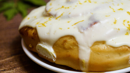 Freshly baked cinnamon roll, generously topped with rich cream cheese frosting and zesty lemon, resting on a white plate atop a rustic wooden table, inviting indulgence and comfort