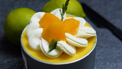Small mango mousse cake is garnished with whipped cream rosettes, fresh mango pieces, and a sprig of cilantro, sitting on a dark gray surface with whole limes nearby