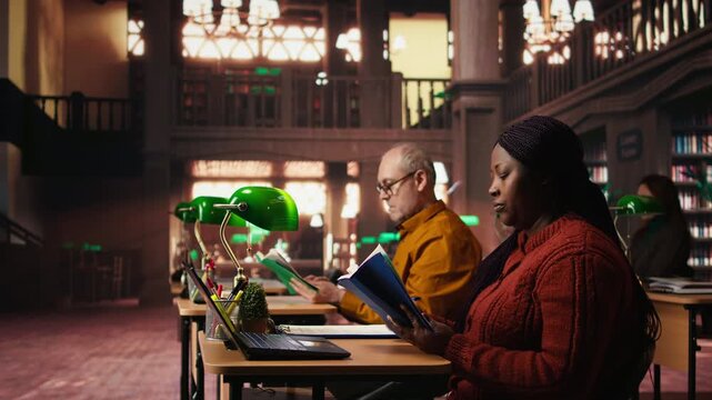 Black female student taking notes surrounded by books and study materials, preparing for a master dissertation and researching academic resources at the campus library. Camera A.