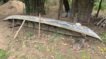 Kracheh, Cambodia - 29 June 2025. A weathered wooden boat lies upside down on the ground beside a bamboo shelter, supported by logs and leaning poles.