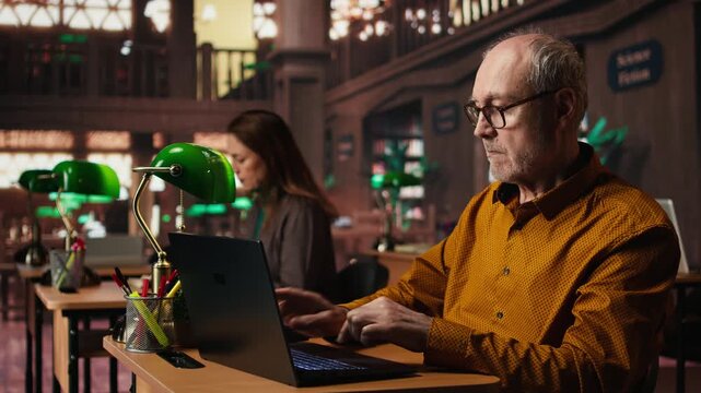 Focused senior guy reading academic text on laptop at the university, sitting in vintage study room and browsing online documents. Lifelong education through formal learning. Camera A. - Powered by Adobe