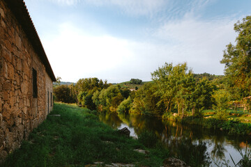 Fototapeta premium Calm waters of the Varosa River reflecting the surrounding lush vegetation and an old stone watermill in Ucanha, Tarouca, Viseu district, Portugal, creating a peaceful rural landscape