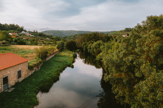 River Varosa flowing calmly through the municipality of Ucanha, Tarouca, with lush vegetation on the riverbanks and a traditional stone house on a cloudy day in Viseu, Portugal