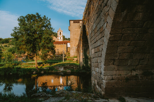 Warm sunlight illuminating a historic stone bridge arching over a tranquil river in Ucanha, Tarouca, Portugal, with a charming village and church in the background