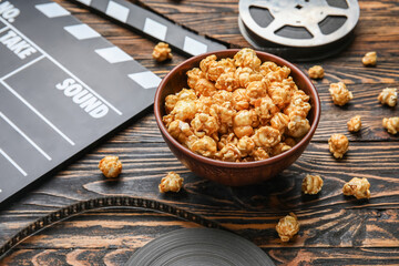Bowl with tasty popcorn, clapperboard and film reel on wooden background