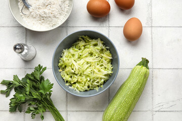 Bowls with grated zucchini, flour and eggs for preparing fritters on white tile background