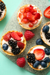 Board of tasty tartlets with whipped cream and berries on turquoise background