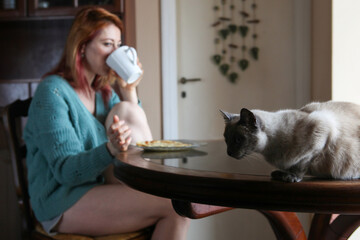 Cozy indoor scene with ginger-haired woman, 30s, sitting at table in home wear. Having breakfast...
