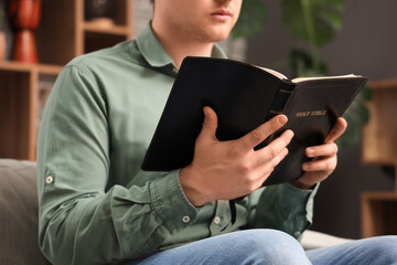 Young man with Holy Bible praying on sofa at home, closeup