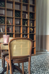 Cozy and stylish interior space featuring a wooden dining table with rattan-back chairs in the foreground, set against a large decorative shelf filled with eclectic vintage items and books