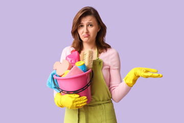 Obraz premium Upset young woman with bucket of cleaning supplies on lilac background