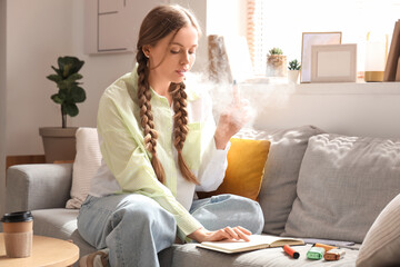 Young woman smoking disposable electronic cigarette and reading book on sofa at home