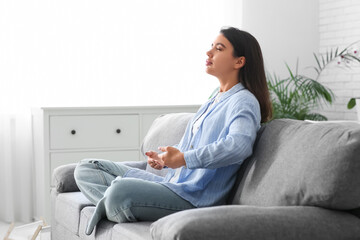 Fototapeta premium Beautiful young woman doing breath practice and meditating on sofa at home