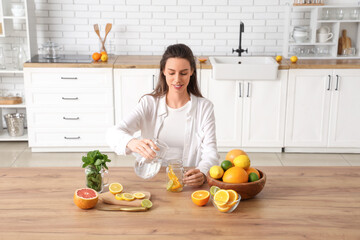 Beautiful young woman preparing infused water with different citrus fruits in kitchen