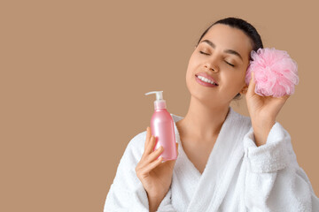 Beautiful young happy woman in bathrobe with bottle of shampoo and sponge on beige background