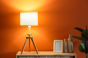 Glowing lamp with blank frame and decor on shelf near orange wall
