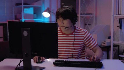 Young Asian gamer programmer student focused deep in concentration playing computer games coding software development wearing headset mic in dark neon lit modern room - Powered by Adobe
