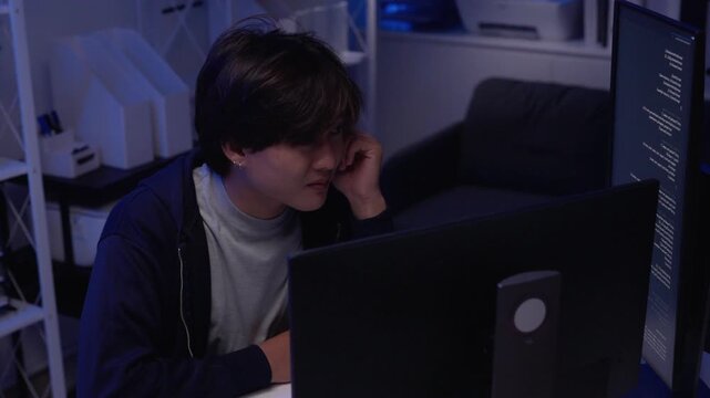 Young Asian adult programmer student intently focused on coding developing complex software solutions late night in a dedicated modern home office setup