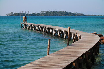 Curved wooden pier extending into turquoise waters with a distant of couple and island backdrop in...